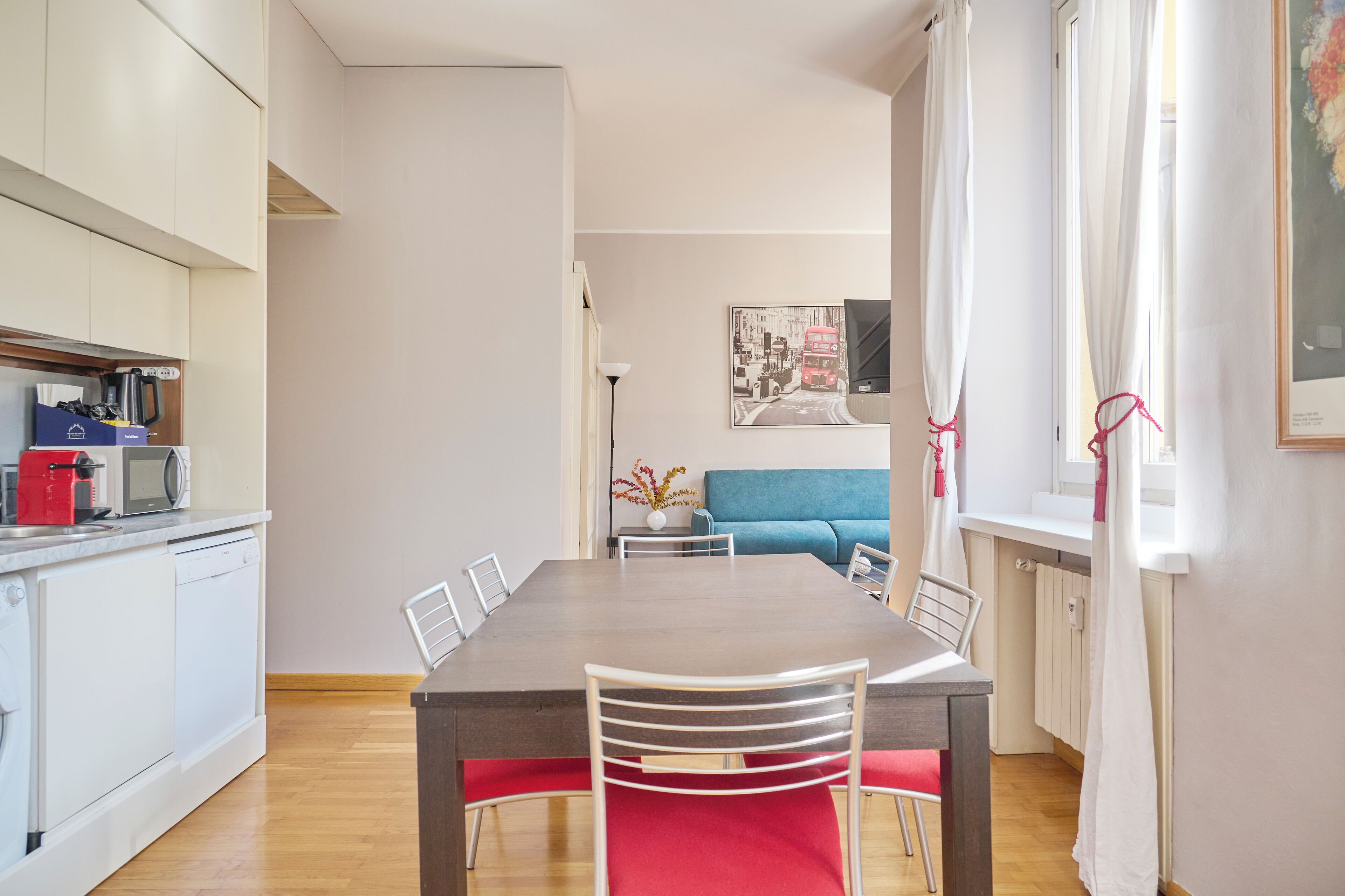 A modern kitchen with white cabinets, a microwave, and a red coffee machine. There is a dining area with a wooden table and white chairs, and a blue sofa in the background.