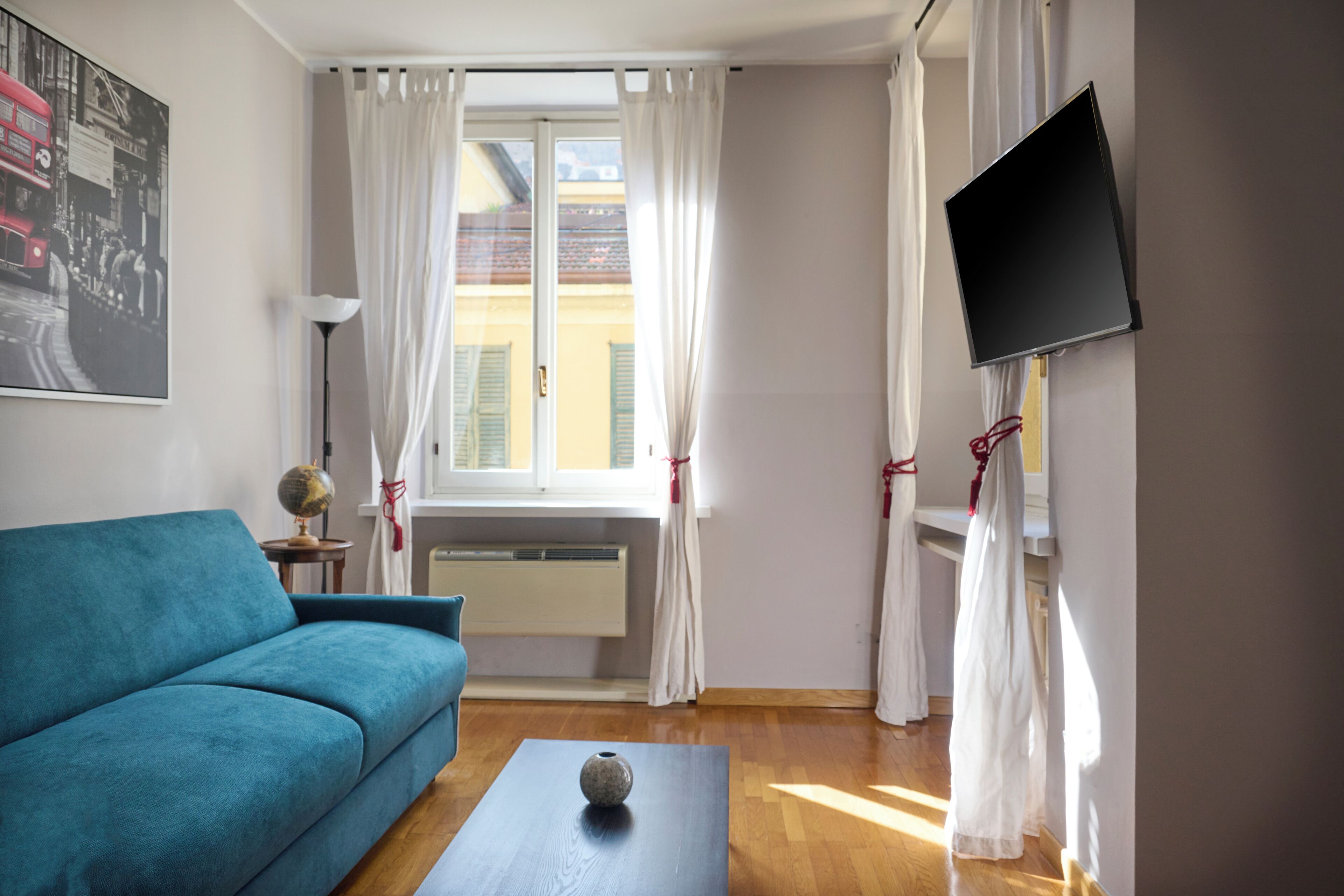 A room with a blue sofa, a small table with a vase, a flat-screen TV mounted on the wall, and a window with white curtains.
