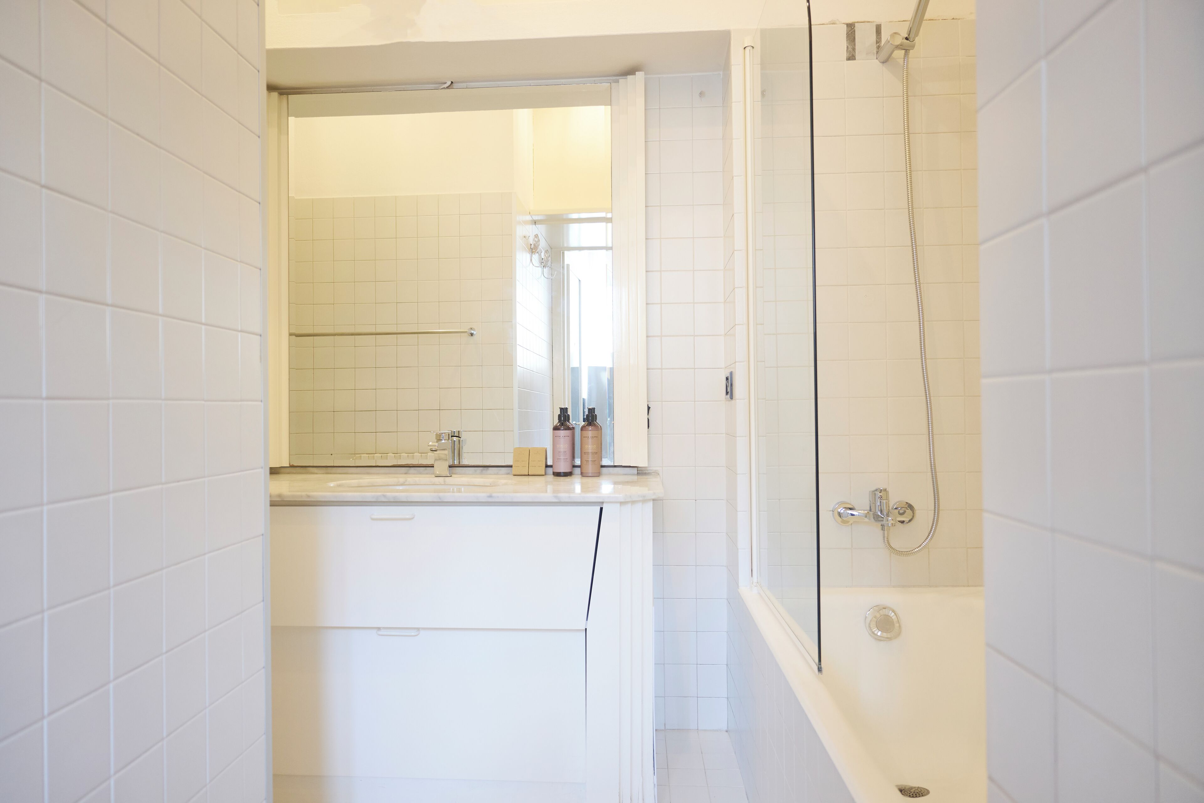 Baño con azulejos blancos, bañera, ducha, lavabo con artículos de aseo y espejo.