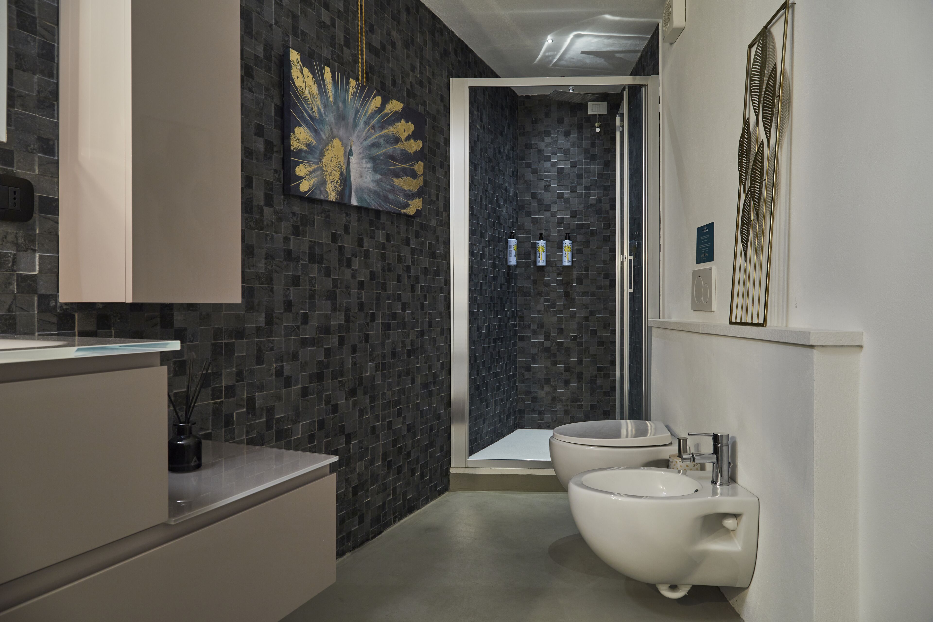 A modern bathroom with a dark mosaic wall, a white sink, and a separate toilet area.