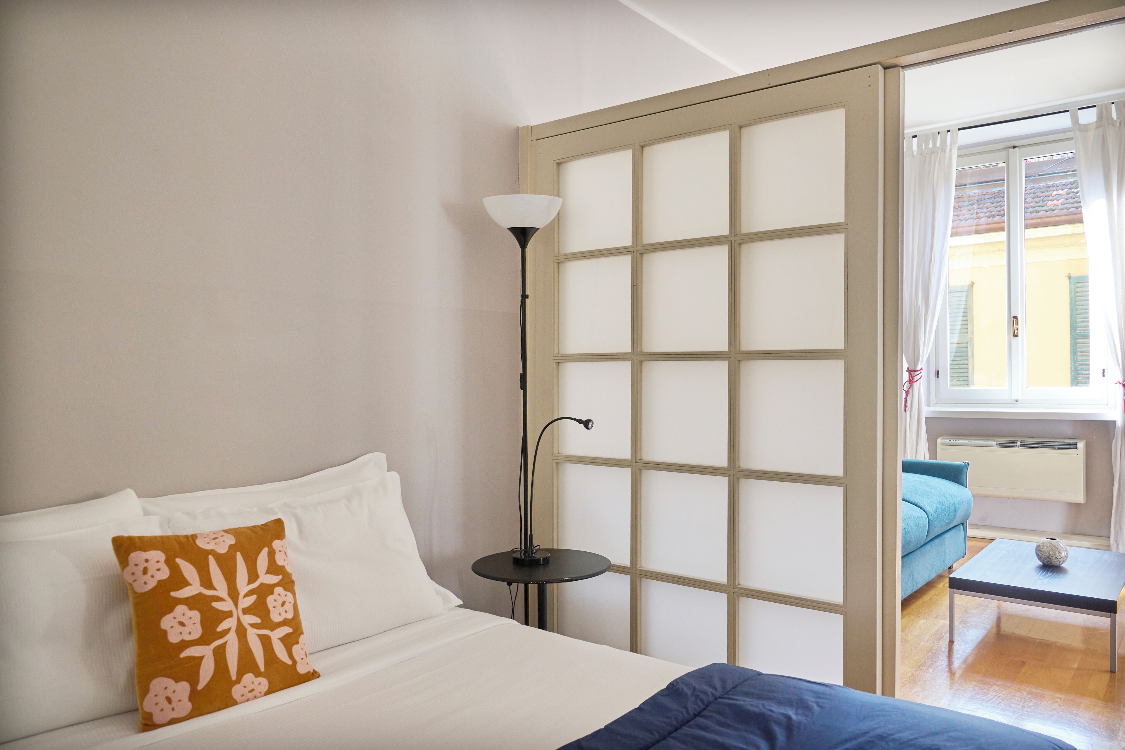 Quarto com cama, uma mesinha, um abajur e vista para área externa.