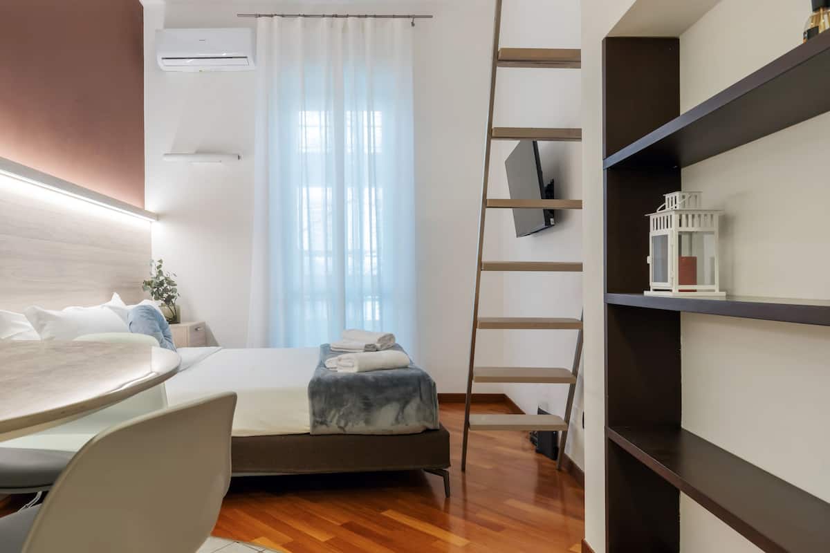 A modern hotel room with a bed, a desk, a chair, a TV, and a bookshelf.