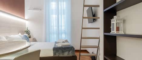 A modern hotel room with a bed, a desk, a chair, a TV, and a bookshelf.