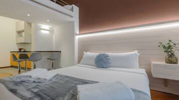 A modern hotel room with a large bed, a bedside table, a mirror, and a kitchenette area with bar stools.