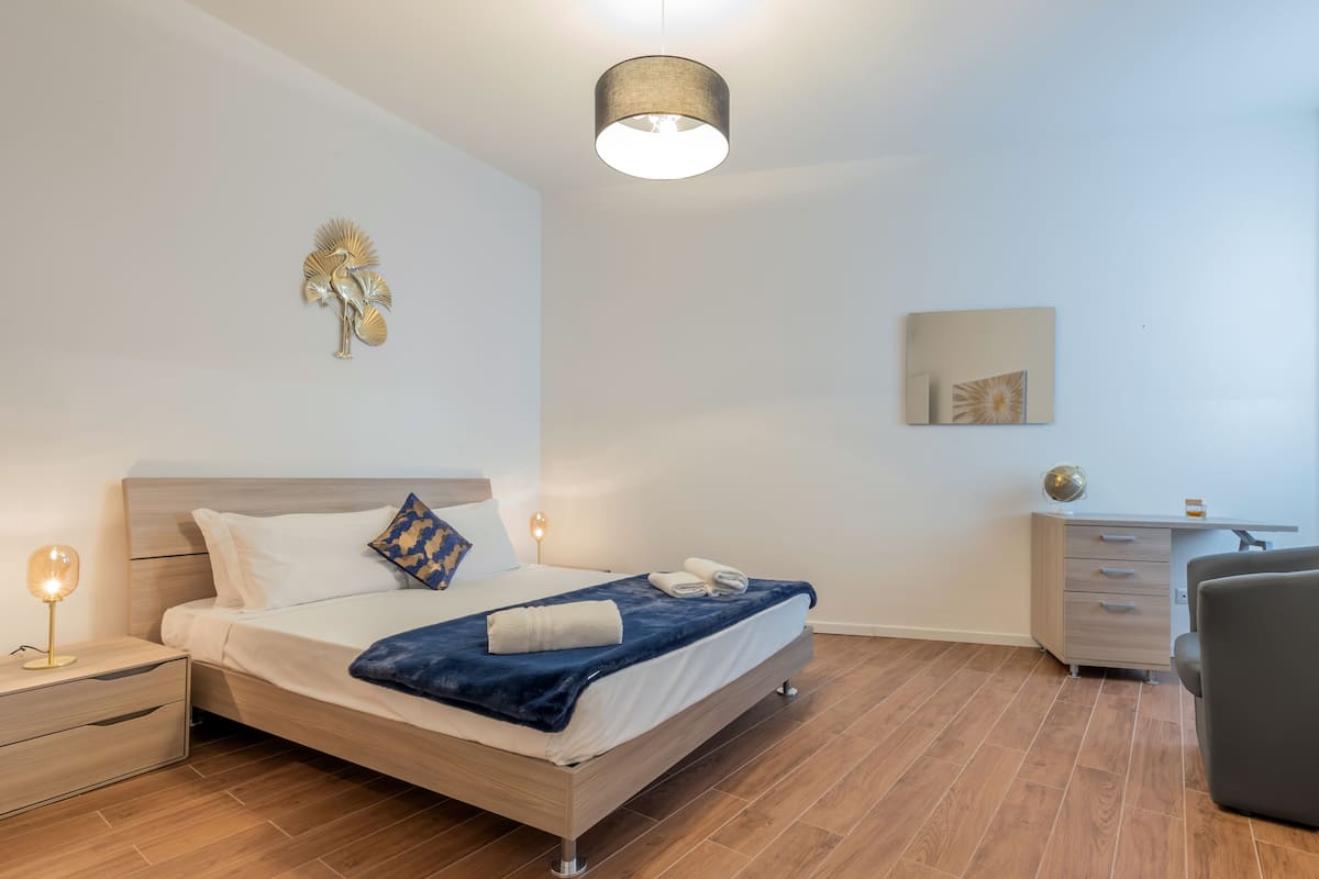 A modern bedroom with a wooden bed, bedside lamps, a desk, and a chair.