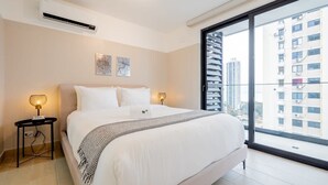Ein modernes Schlafzimmer mit einem großen Bett, zwei Nachttischlampen, einer Klimaanlage und Blick auf eine Stadtlandschaft durch ein großes Fenster.