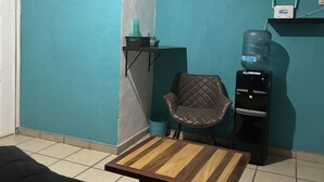 Habitación con paredes color turquesa, un sofá negro, una mesa de centro de madera, un dispensador de agua, una silla y un estante pequeño con un letrero de 'Welcome'.
