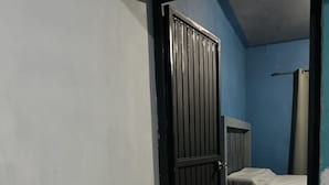 Un estrecho pasillo con una puerta negra cerrada, una cama con cabecera, una mesita de noche y una cortina.