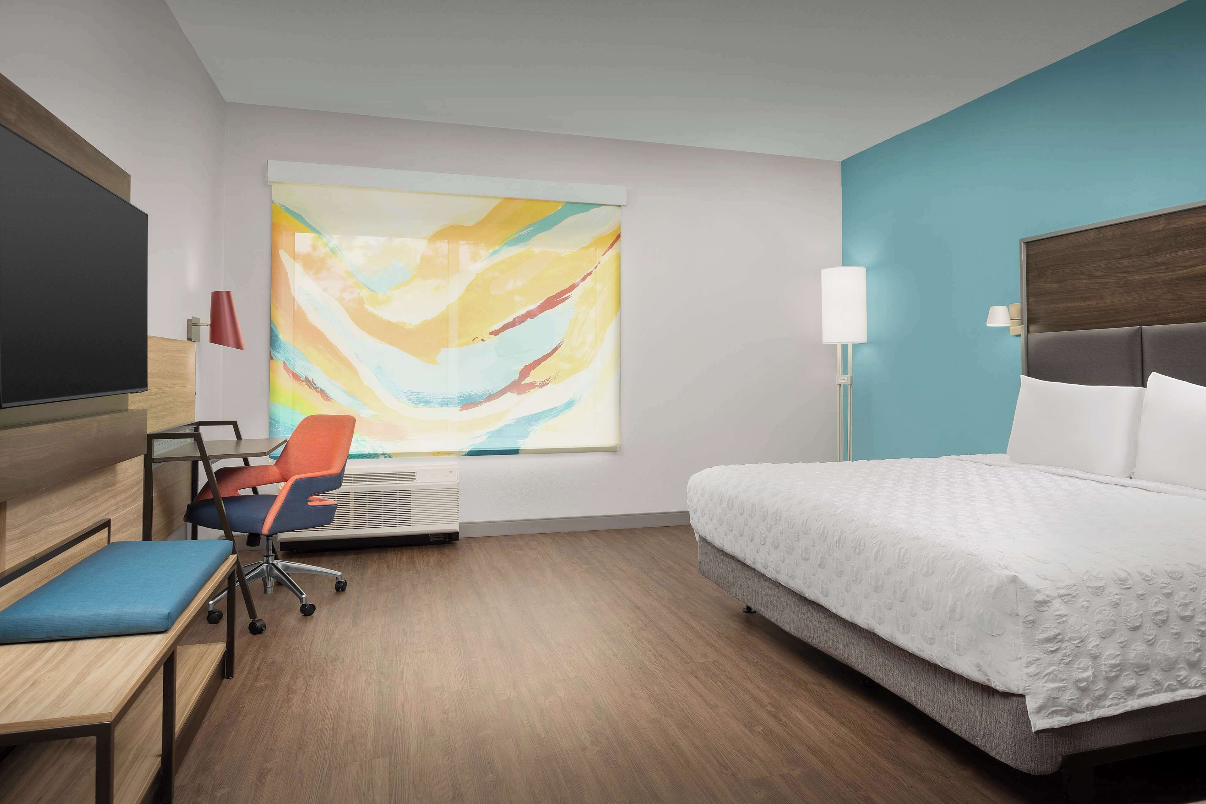 A modern hotel room with a large bed, a desk with a chair, a television, and a colorful abstract painting on the wall.