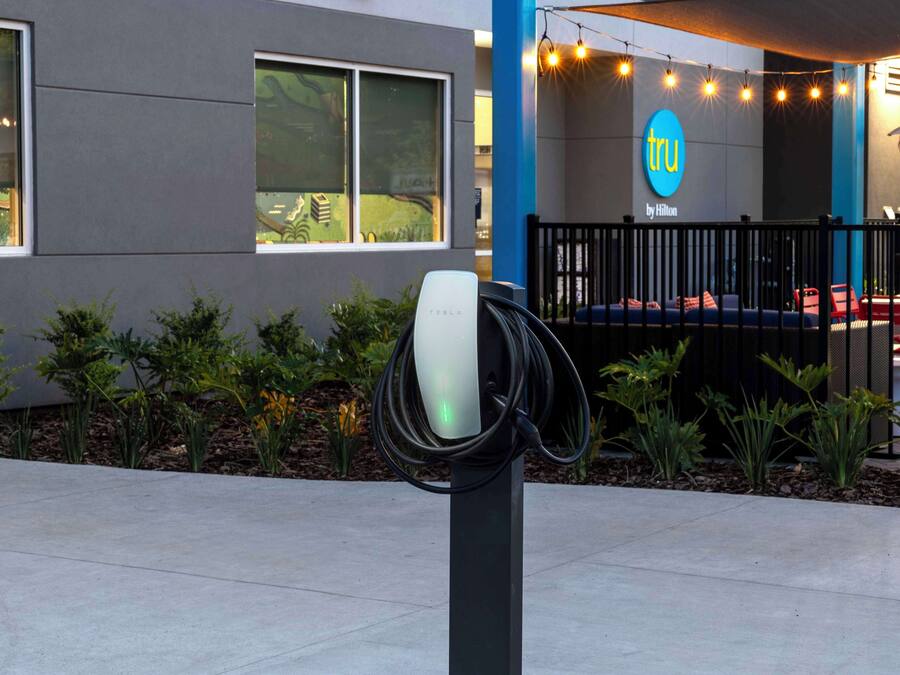 An electric vehicle charging station in front of a modern building with a blue and gray facade, a sign reading "tru by Hilton," and outdoor seating with string lights.