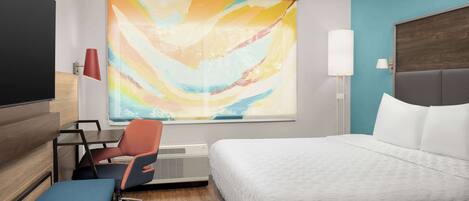 A hotel room with a bed, a desk with a chair, a window with a colorful abstract painting, and a wall-mounted lamp.
