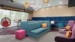 A modern lounge area with a blue sofa, a red chair, and a purple armchair. There are hanging white and orange pendant lights, and a large window providing natural light. - Tru by Hilton Jacksonville West I 295 (Jacksonville)