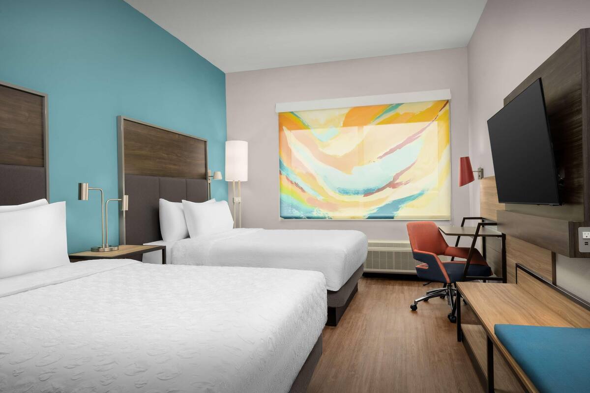 Habitación de hotel con dos camas, un escritorio, una silla, un televisor y un cuadro abstracto grande en la pared.