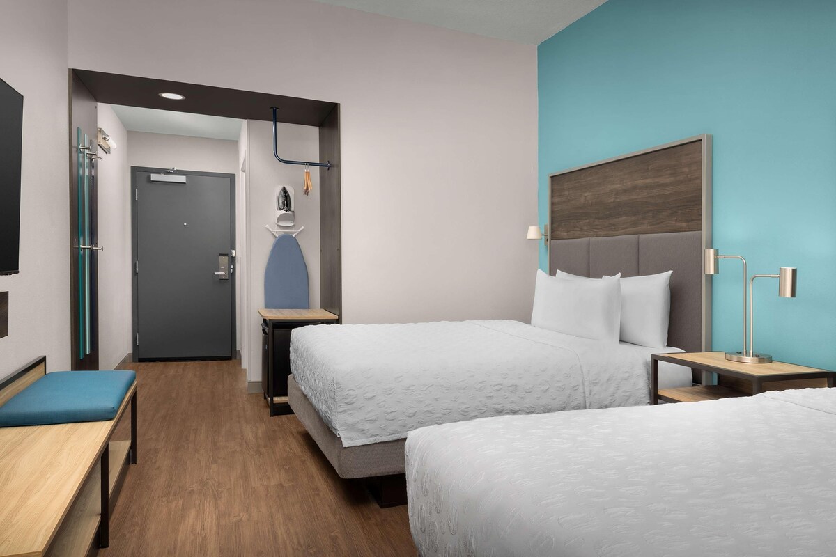 A hotel room with two beds, a desk, a bench, a door, and a wall-mounted TV.