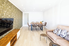 A modern living room with a wooden TV stand, a beige sofa with patterned cushions, a dining area with wicker chairs and a table set with fruits, and a large mirror reflecting the room.