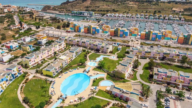 A coastal resort with colorful buildings, a marina, and a large swimming pool.