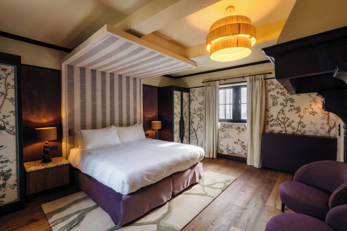 A bedroom with a large bed, a nightstand with a lamp, a wooden floor, and a ceiling light fixture.