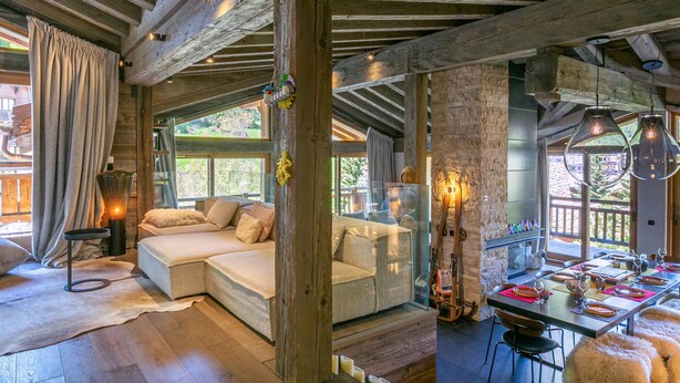 Un interior rústico con vigas de madera a la vista, una zona de estar acogedora con cojines, una mesa de comedor dispuesta para una comida, y un ventanal con vistas al exterior.