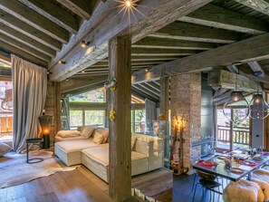 A rustic interior with exposed wooden beams, a cozy seating area with cushions, a dining table set for a meal, and a large window offering a view of the outdoors. - Stunning Top-Floor 5-Bedroom Apartment with Panoramic Views Near Slopes (Val-d'Isère)