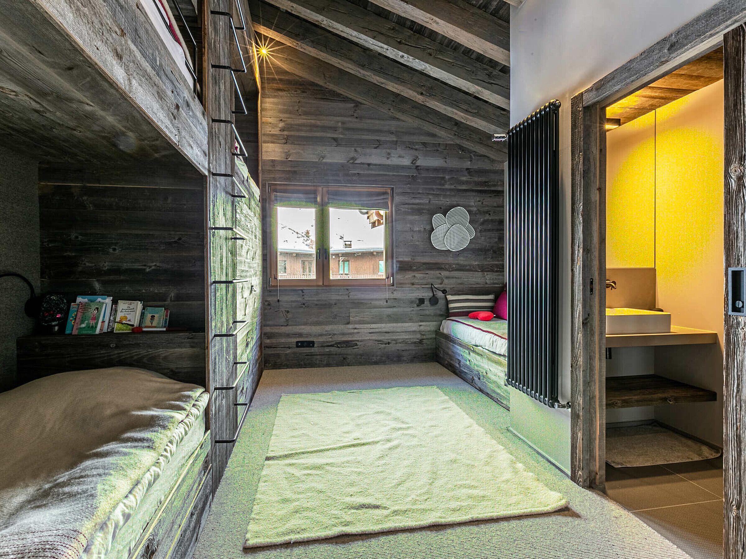 Een eenvoudige houten kamer met een houten plafond, een raam met uitzicht, een bed met een groene matrras en een badkamer met een wastafel en spiegel.