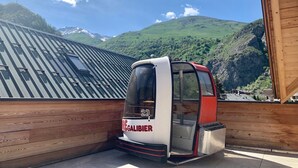 A small, enclosed vehicle labeled "REGALIBIER" parked on a wooden deck with a view of green hills and a mountain range in the background. - 4* Chalet for 8, sunny with wifi and pets allowed (Valloire)