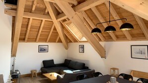 A cozy loft-style room with exposed wooden beams, a dark sofa, a dining table, and a small kitchen area. - 4* Chalet for 8, sunny with wifi and pets allowed (Valloire)
