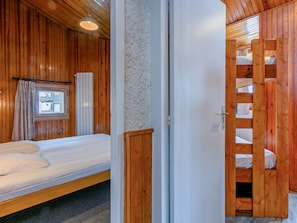 Une pièce de style cabane en bois, avec un lit simple, une fenêtre avec des rideaux et une porte en bois donnant sur une mezzanine équipée de lits superposés.