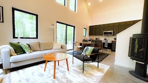A spacious living area with a sofa, chairs, a coffee table, and a fireplace. A kitchen is visible in the background with modern appliances. - Hodag House | Magical Cabin & Creature Comforts (Herbster)