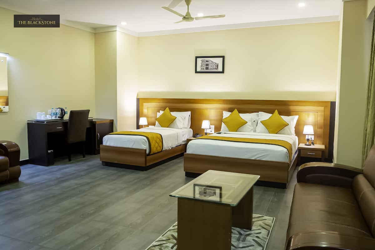 A hotel room with two beds, a desk, a chair, a sofa, and a ceiling fan.