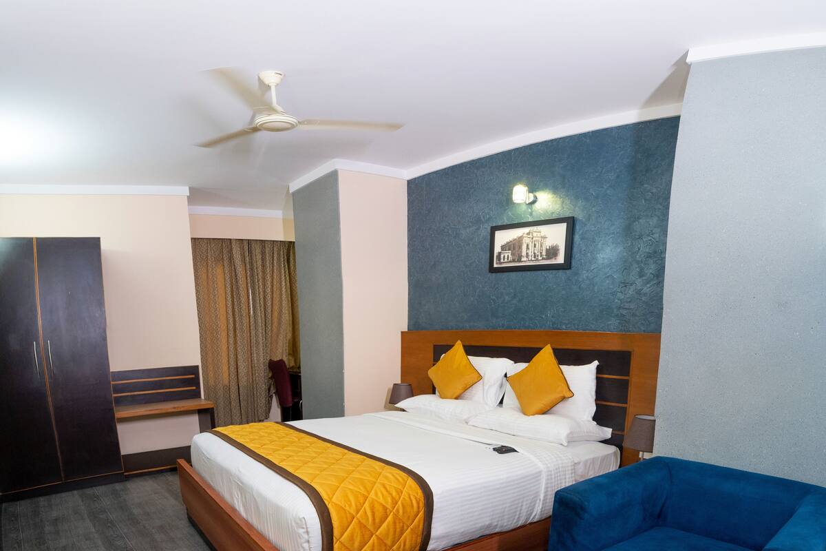 A hotel room with a bed, a blue armchair, a wooden coffee table, a ceiling fan, and a framed picture on the wall.