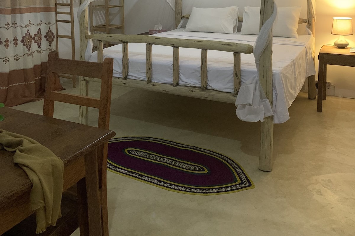 A bedroom with a wooden four-poster bed, a wooden chair, a small table, and a nightstand with a lamp.