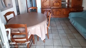 A dining area with a round table, chairs, a cabinet with glass doors, and a bed with a blue cover. - Petite Maison Avec 2 Chambres 3 Personnes Avec Terrasse Propriété Fermée (Calvi)
