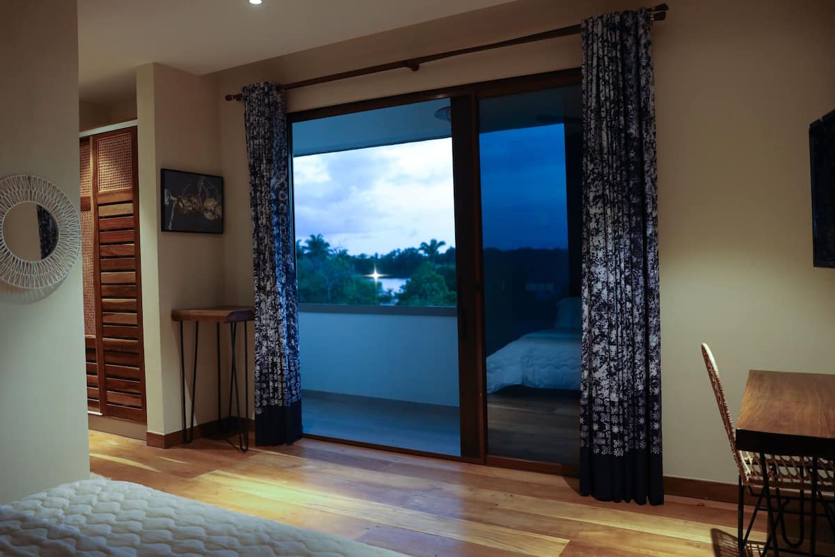 A bedroom with a wooden floor, a bed with a white duvet, a small table, a chair, a window with curtains, and a view of a landscape.