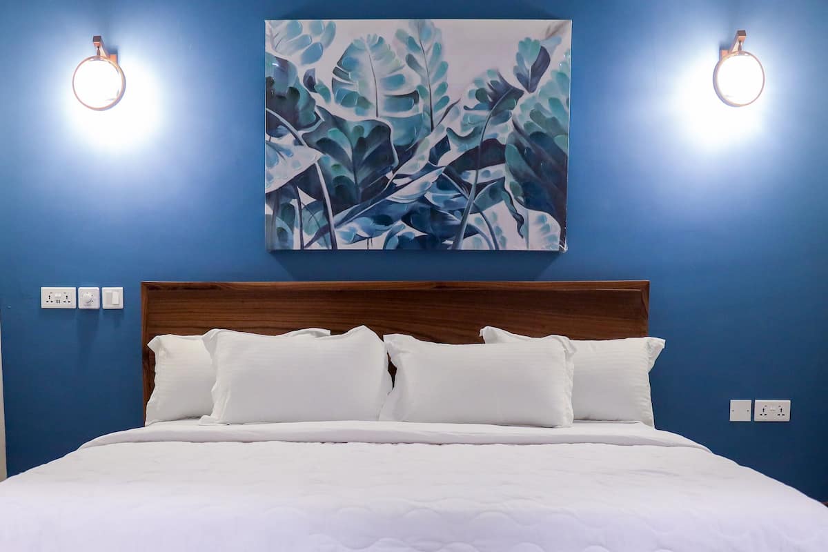 A bedroom with a large bed, a wooden headboard, and a blue wall with a leaf-patterned painting. There are two wall-mounted lights and a power outlet on the wall.