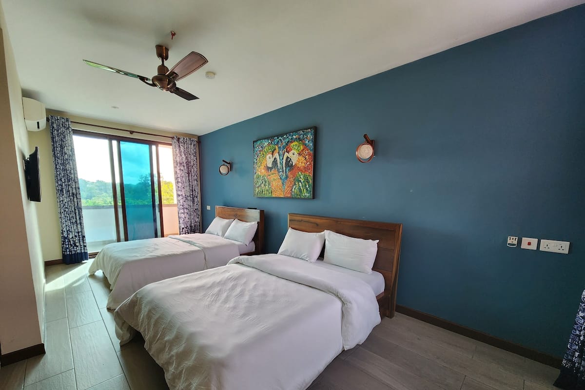 A hotel room with two beds, a ceiling fan, a painting, and a view of the outdoors.