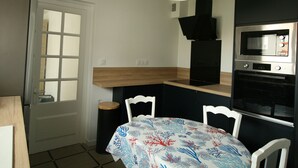 A compact kitchen with a white door, a wooden countertop, a black range hood, a microwave, and a table with a floral tablecloth. - Charming House Near Beach and Shops with Private Parking and Terrace (La Tranche-sur-Mer)