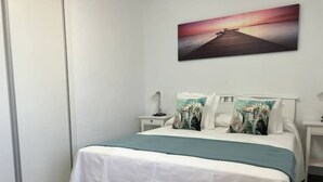 A bedroom with a bed, white bedding, a nightstand with a lamp, and a wall-mounted painting of a sunset over a pier.
