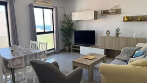 A modern living room with a dining area, a flat-screen TV, a sofa, and a balcony with a view.