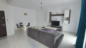 A modern living room with a grey sofa, a dining table with chairs, a TV, and a kitchen area with cabinets and a microwave. - VISAJ Metropolitan (Giroc)