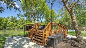 A wooden deck with a staircase leading to a covered area, surrounded by trees and a body of water. - River Bend Bliss: Where Memories flow with the River (Cedar Creek)