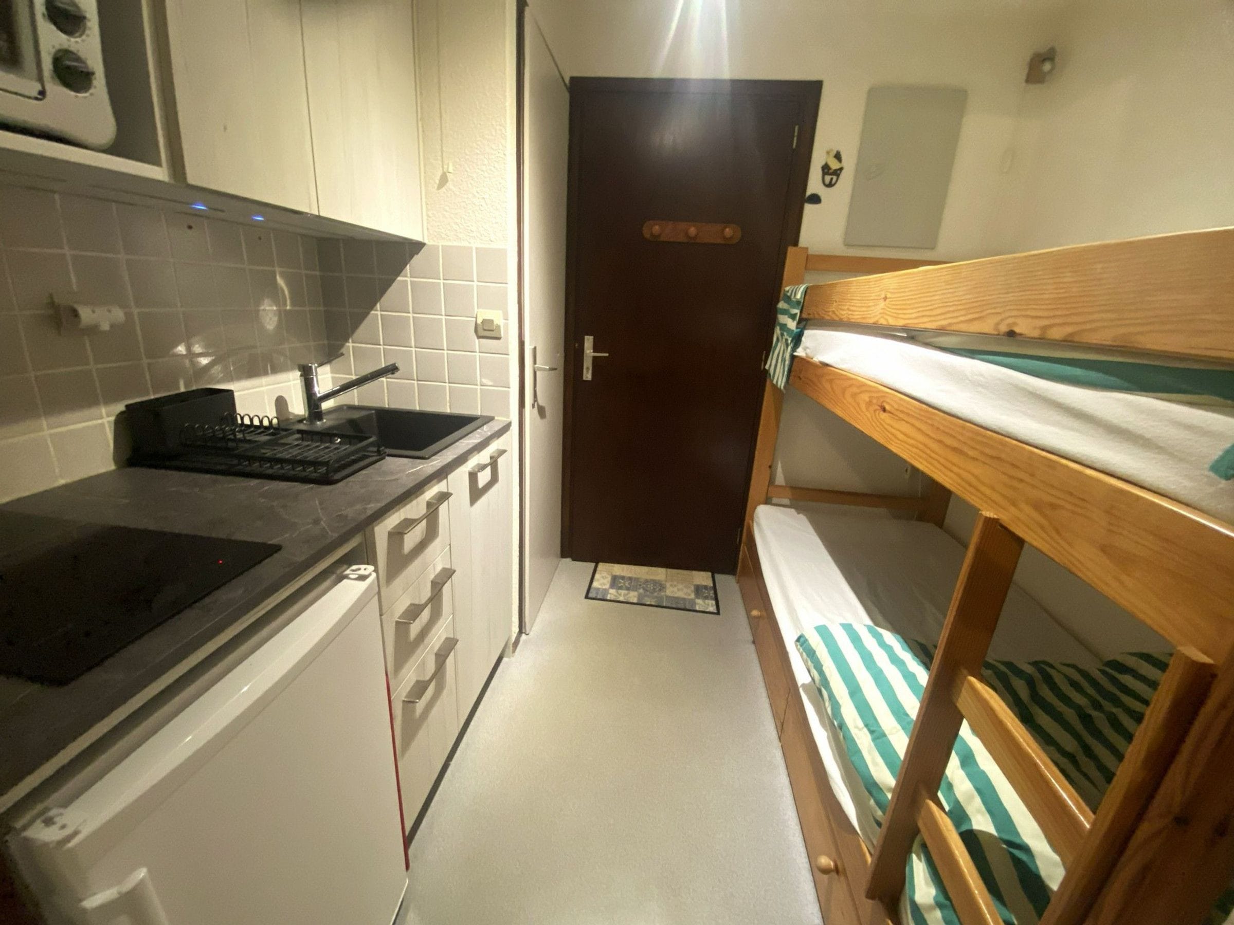A compact kitchen with a sink, stove, and cabinets, a wooden bunk bed with striped bedding, and a door with a handle.