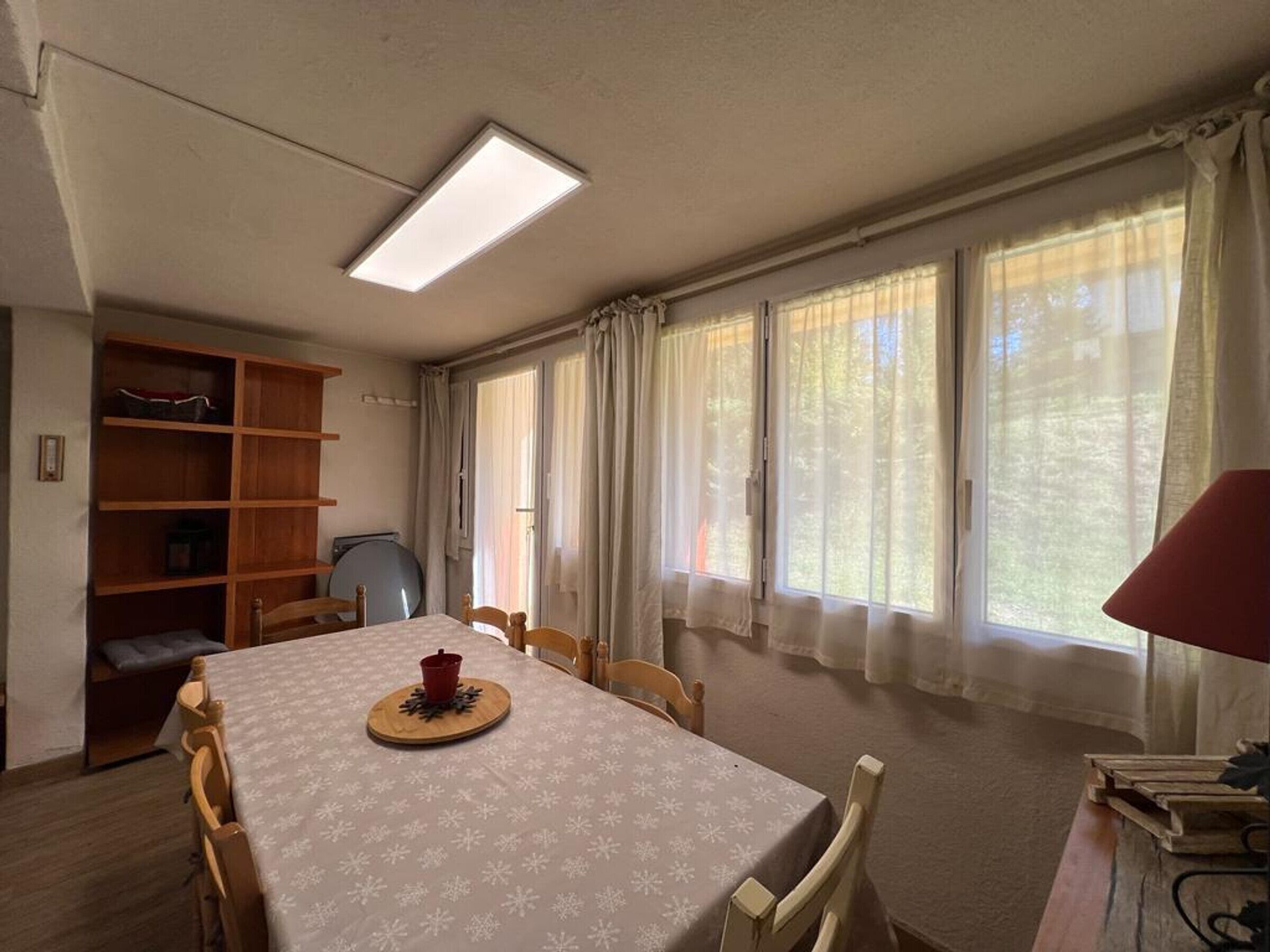 A room with a dining table, chairs, a bookshelf, and large windows with curtains.