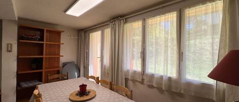 A room with a dining table, chairs, a bookshelf, and large windows with curtains.