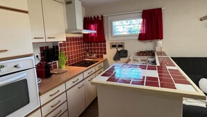 A compact kitchen with white cabinets, a red-tiled backsplash, a window with red curtains, and a small island with a tiled countertop. - Spacious apartment for 8, garden and WIFI, near shops in Pra Loup (Pra-Loup)