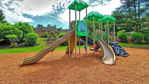 A playground with slides and climbing structures, surrounded by a landscaped garden and a building in the background.
