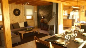 Un intérieur chaleureux en bois, avec un coin repas aménagé pour prendre un repas, un salon doté d’un canapé et d’une cheminée, et une cuisine visible au fond.