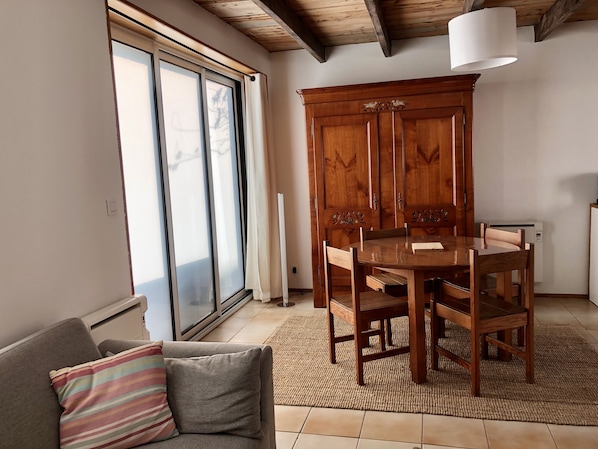 Un coin repas avec une table et des chaises en bois, un meuble orné de sculptures décoratives, un canapé avec un coussin à rayures et une grande fenêtre avec des stores.