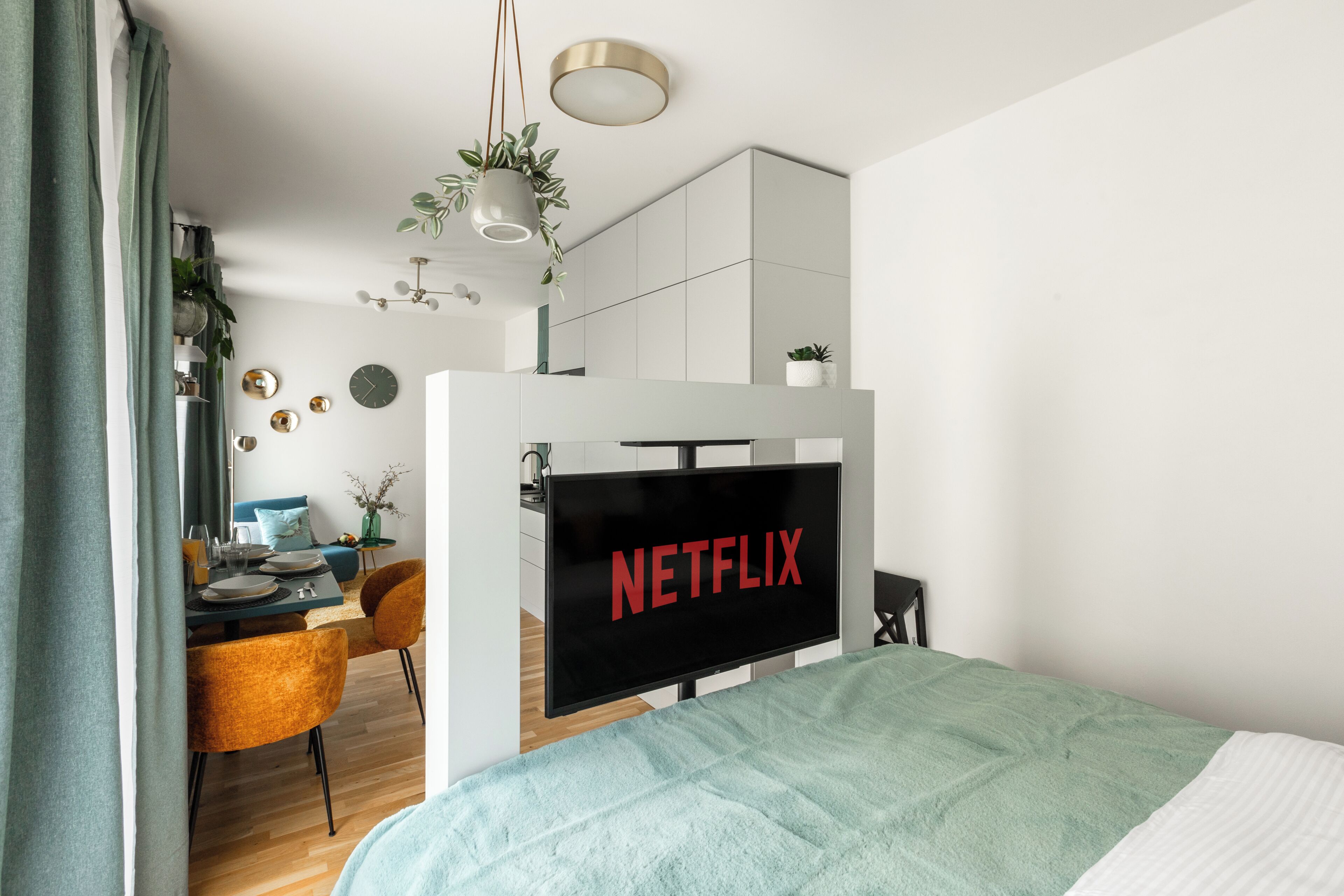 Et moderne soveværelse med seng, et fjernsyn der viser 'NETFLIX', et spiseområde med stole og bord samt et køkkenområde.