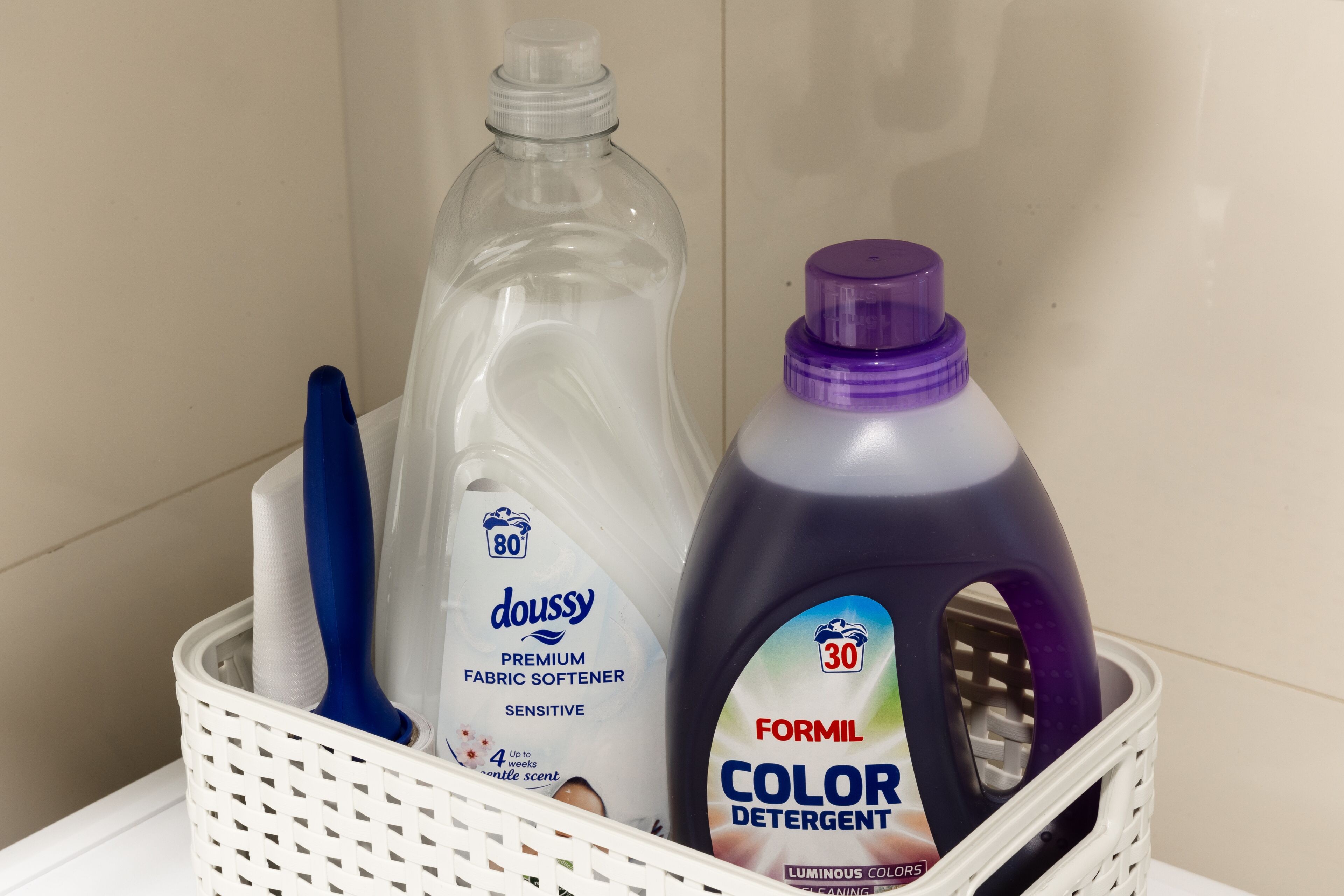 A basket containing a bottle of Doussy fabric softener, a bottle of Formil color detergent, and a blue-handled brush.