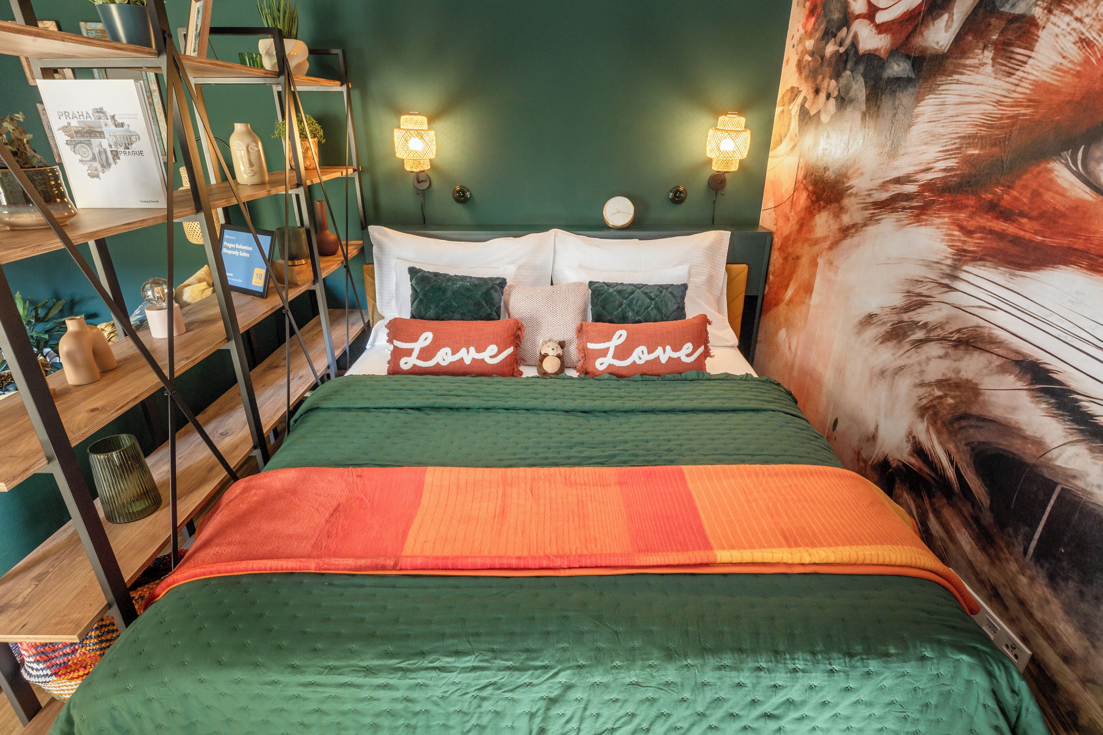Una cama con ropa de cama verde y naranja, dos almohadas con el texto 'Love', un reloj de pared y una estantería con varios objetos.